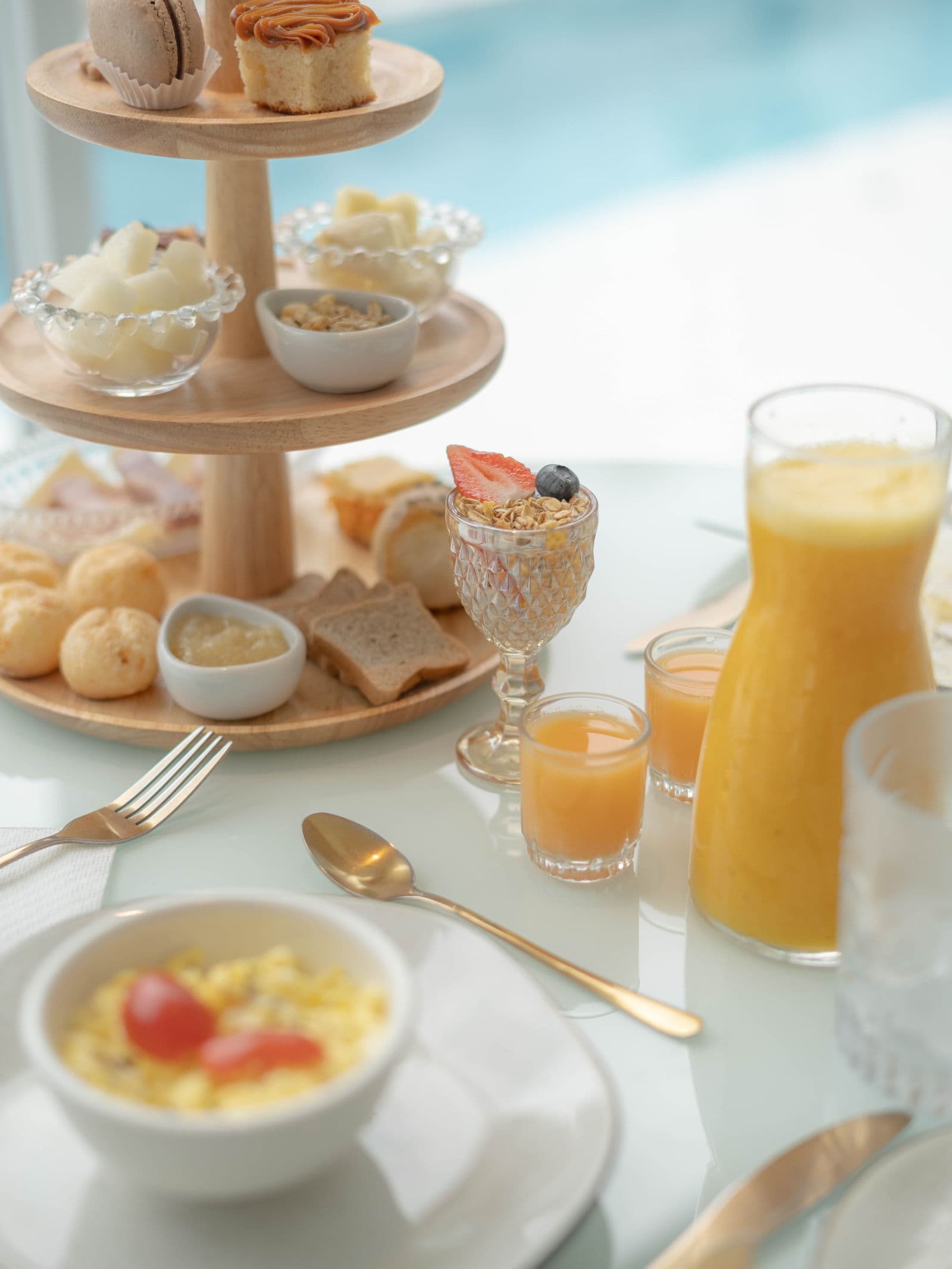 Torre de café da manhã com macarons, bolos e pães de queijo, ao lado de uma jarra de suco de laranja e ovos mexidos