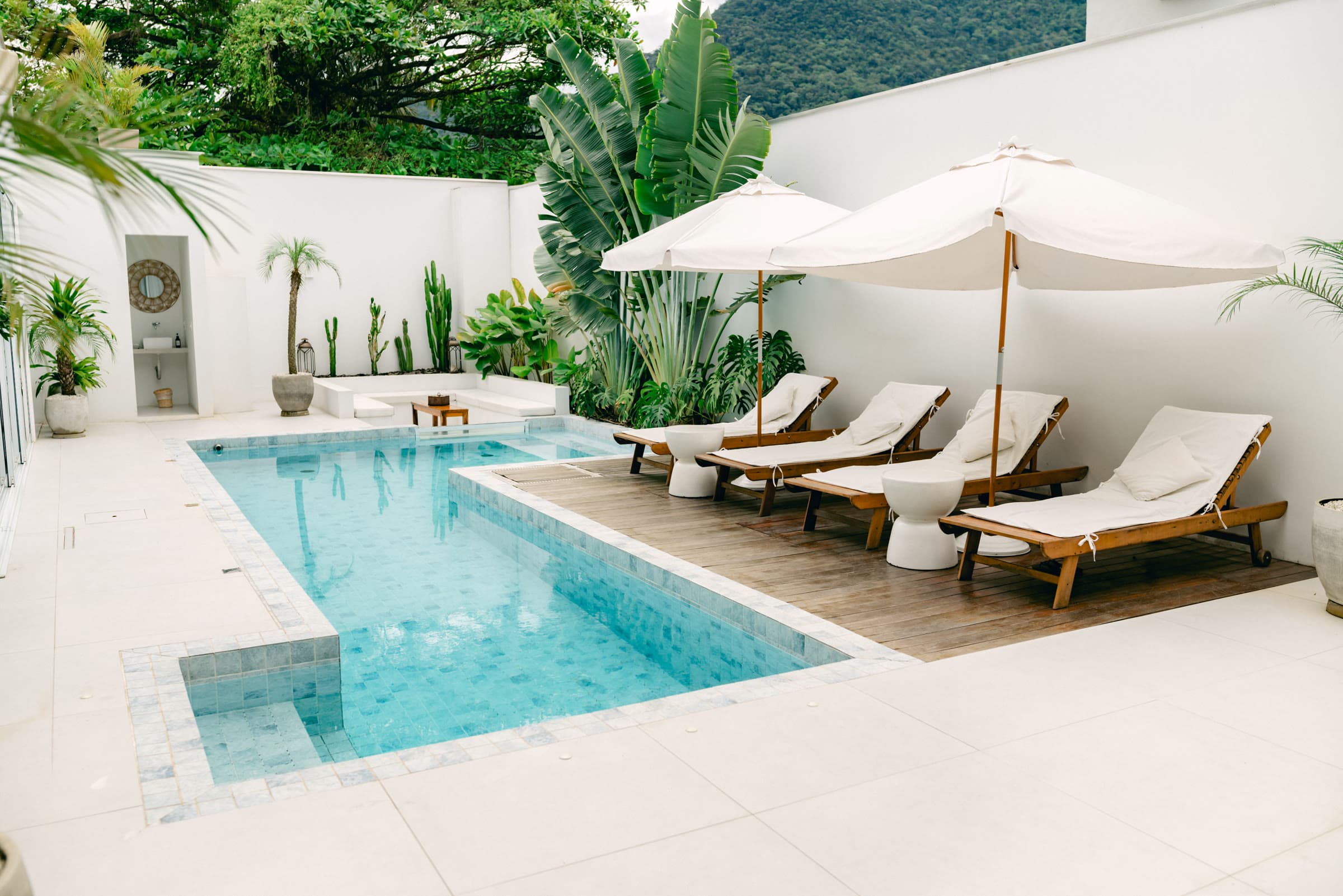 Piscina aquecida do OLISA Boutique Hotel com quatro espreguiçadeiras de madeira sob dois guarda-sóis brancos no deck, cercada por bananeiras, cactos e palmeiras, com a Mata Atlântica atrás do muro caiado