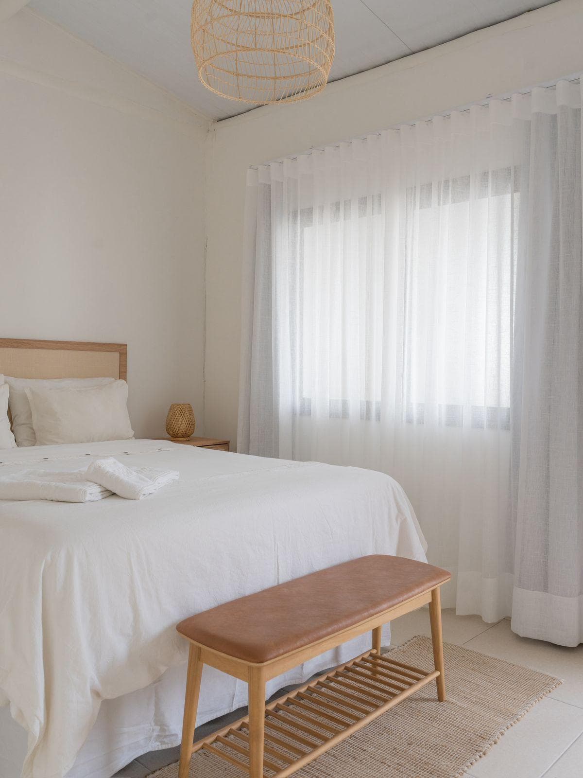 Cama de casal com cabeceira de palhinha e roupa de cama de linho branco, banco de madeira e couro ao pé, pendente de fibra trançada e janela larga com cortina de voil