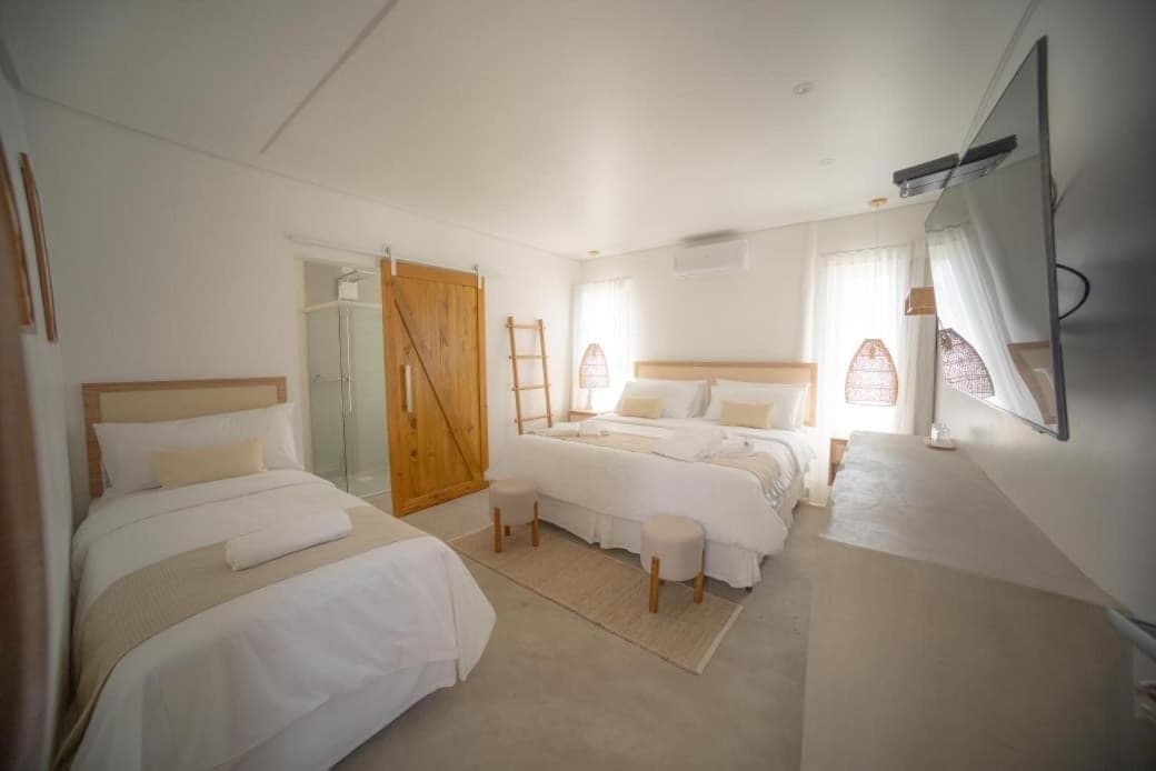 Quarto visto do canto, com a cama de solteiro em primeiro plano e a cama king-size ao fundo, dois pufes redondos sobre o tapete de juta, porta de madeira de correr para o banheiro de box envidraçado, escada decorativa de madeira e televisão de tela plana na parede