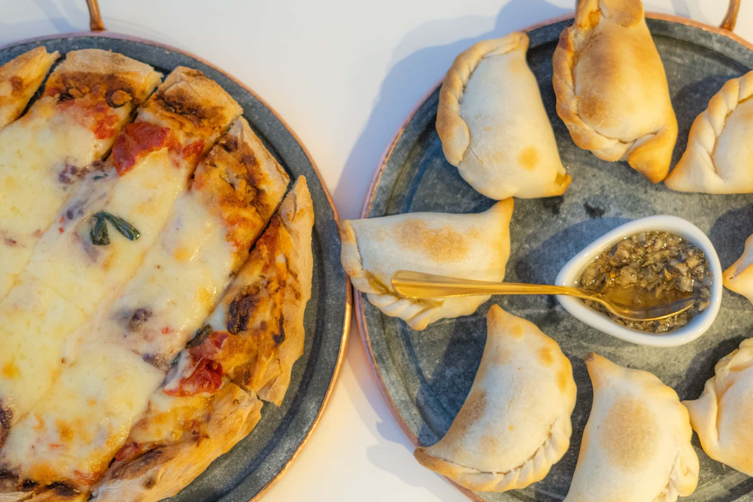 Pizza cortada em fatias e empanadas dispostas em travessas de pedra escura, com um potinho de molho no centro