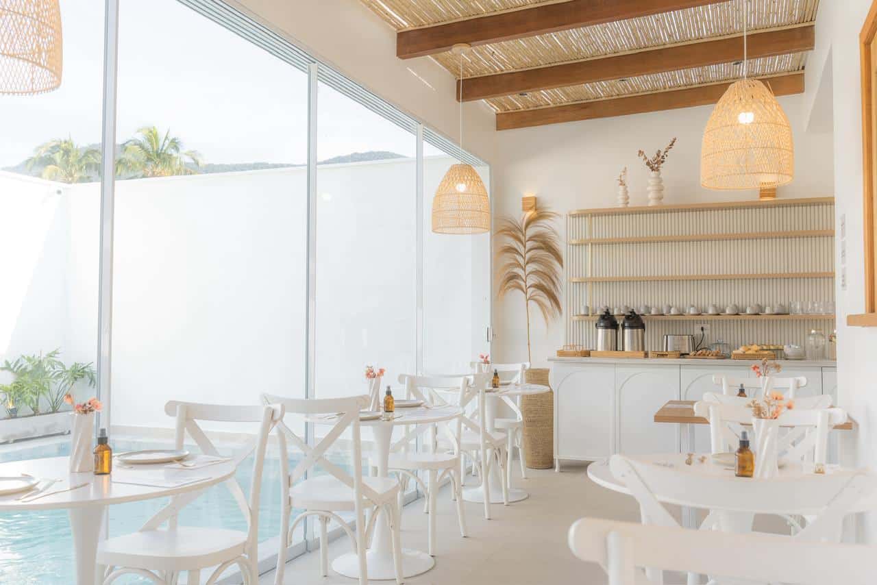 Salão visto de dentro: vidro do chão ao teto com a piscina do outro lado, mesas redondas brancas, cadeiras bistrô, forro de bambu com vigas de madeira e o balcão do buffet ao fundo