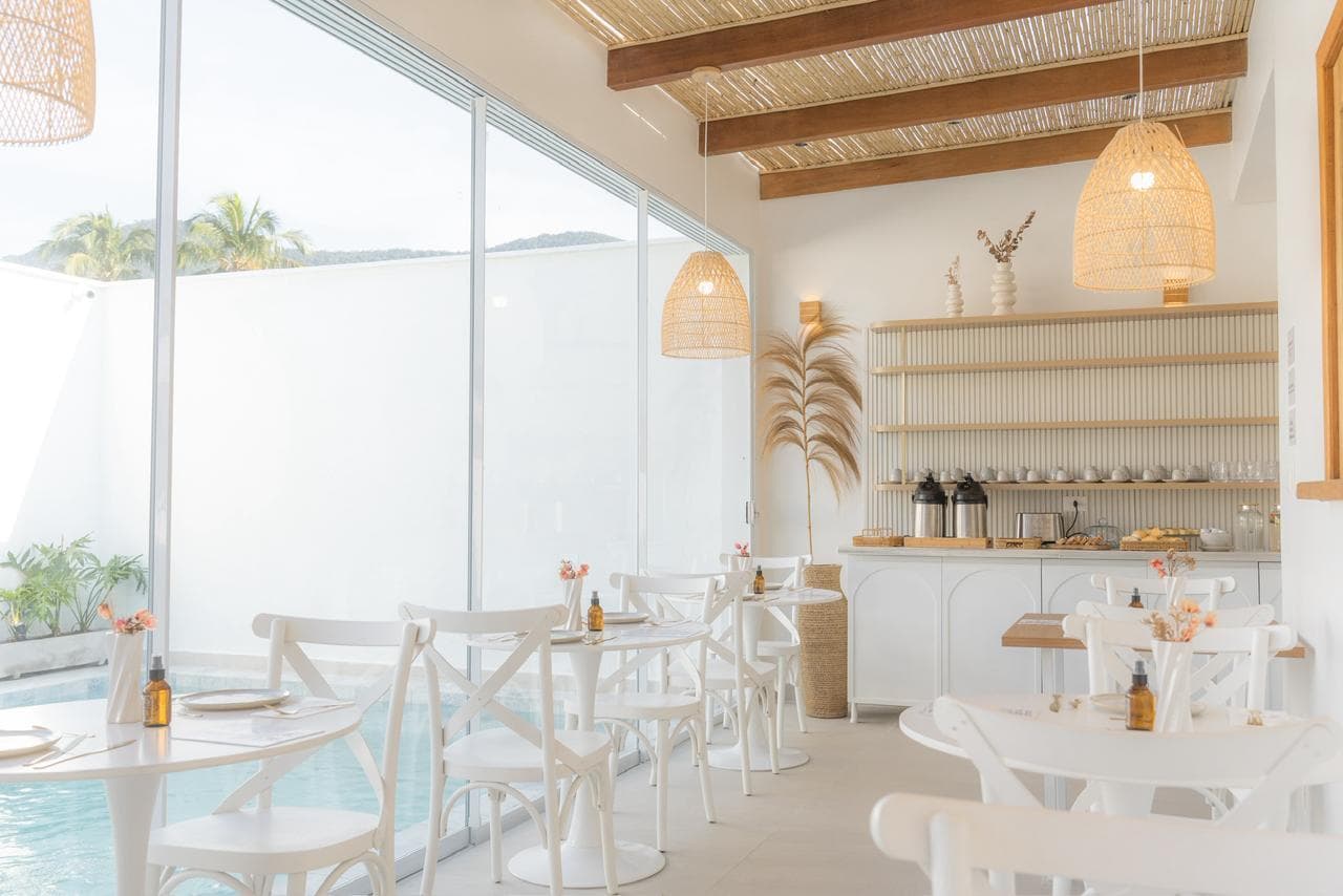 Salão visto de dentro: vidro do chão ao teto com a piscina do outro lado, mesas redondas brancas, cadeiras bistrô, forro de bambu com vigas de madeira e o balcão do buffet ao fundo