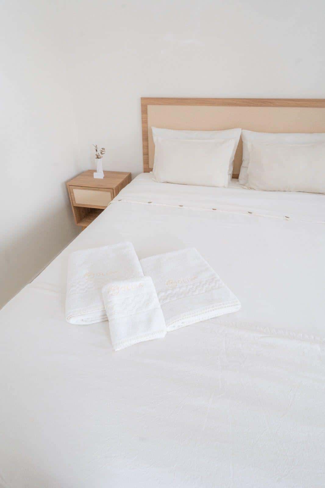 Toalhas brancas com o logotipo OLISA bordado em dourado, dobradas sobre a cama, ao lado da cabeceira de madeira e palhinha e da mesa de cabeceira com uma flor seca