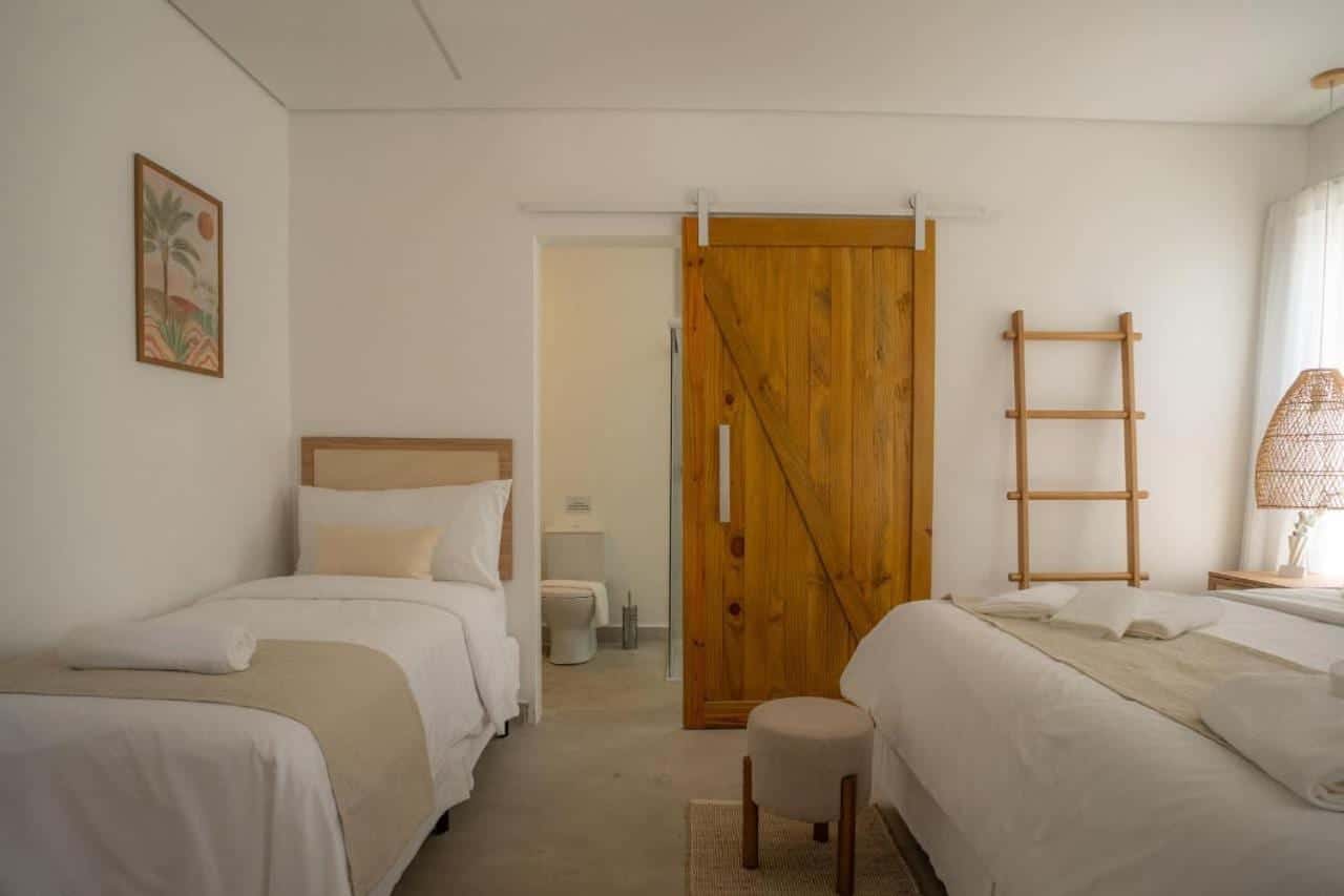 Quarto com a cama de solteiro de um lado e a king-size do outro, a porta de madeira de correr aberta para o banheiro, escada decorativa de madeira, quadro de palmeiras na parede e abajur de fibra trançada
