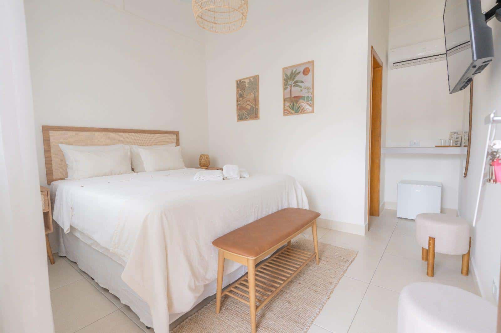 Suíte inteira num quadro só: cama de casal com cabeceira de palhinha, banco de madeira e couro ao pé, tapete de juta, dois quadros botânicos, frigobar, televisão na parede e a porta de madeira do banheiro
